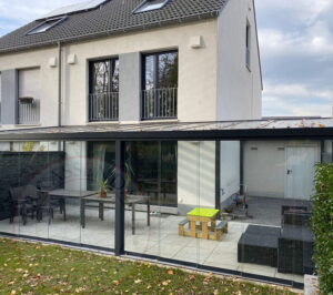 Terrassenüberdachung mit Glasdach und seitlichen Glas-Schiebewänden aus Aluminium an einem Reihenhaus, mit überdachter Terrasse und Gartenmöbeln